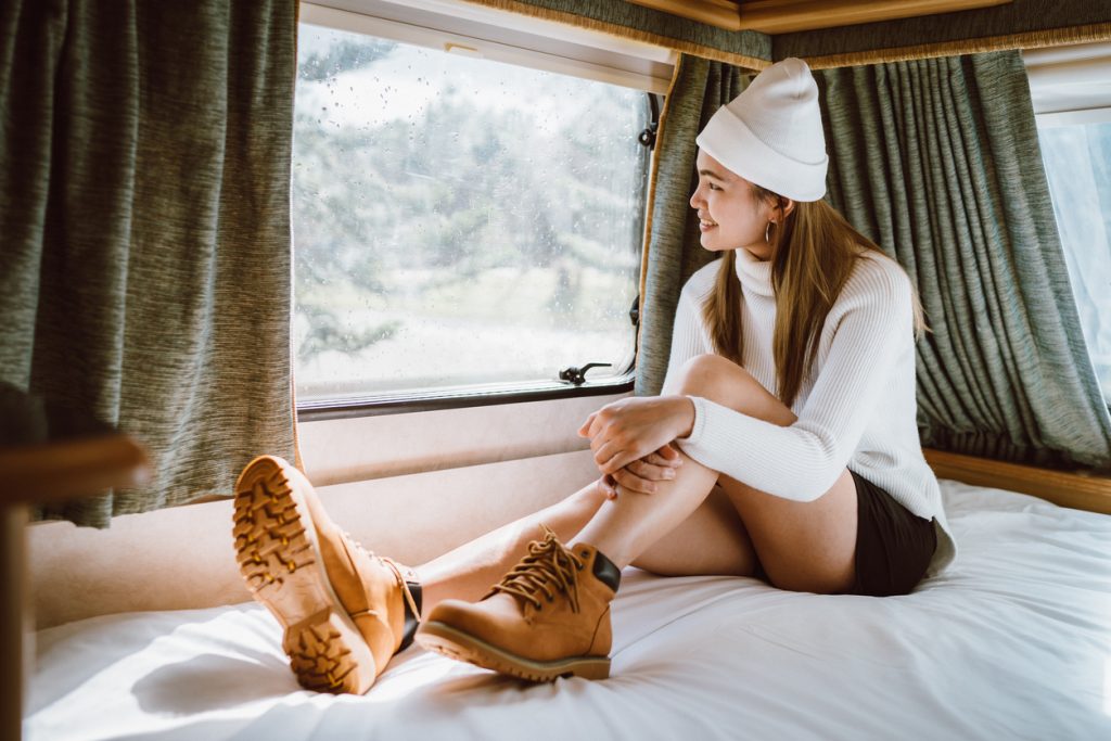 She is sitting on the bed in the Recreational vehicle home car, See the pine forest view outside the window.